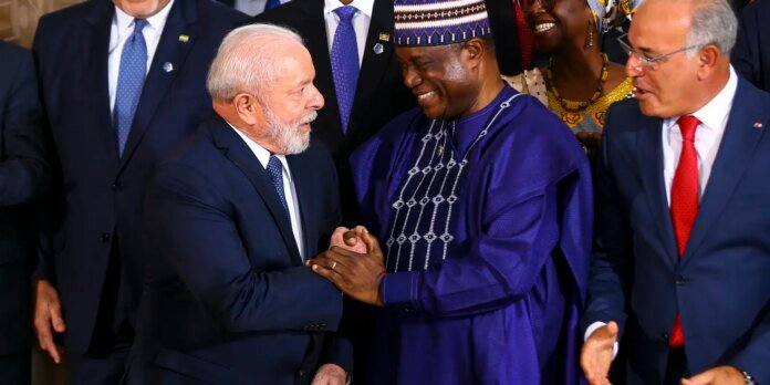 Two men in formal attire shake hands and smile during a group gathering, others in suits and colorful traditional clothing nearby