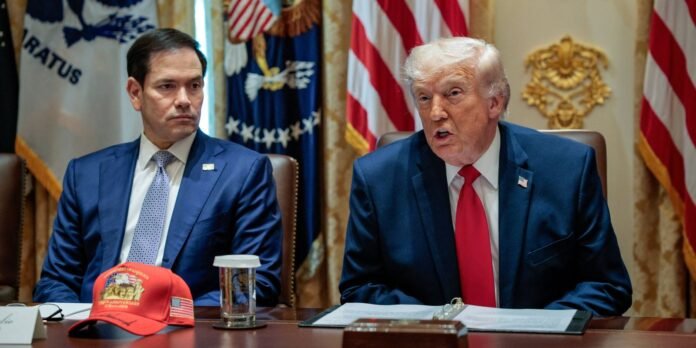 Two men in dark suits sit at a conference table with documents, a red cap, and a glass of water in front, flags and gold decor in the background.