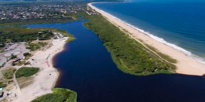 Aerial view of a coastal area: sandy beach along the ocean on the right, a narrow river runs parallel with green vegetation in between, and a town with houses inland on the left.