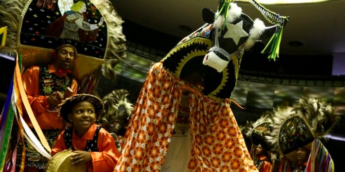 Group of performers in bright costumes dancing around a large decorated horse head prop.
