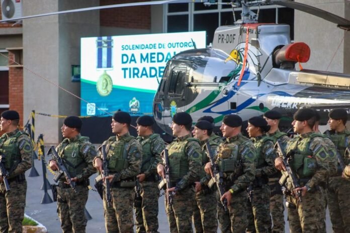 Line of soldiers in camouflage uniforms with rifles, standing in front of a helicopter during a ceremony.