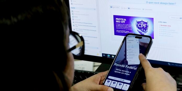 Person holding a smartphone showing a Brazilian federal tax app ('Receita Federal') in front of a computer monitor displaying a related webpage.