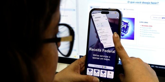 Pessoa segurando um celular na vertical com o app Receita Federal aberto, exibindo opções de serviços; monitor ao fundo com página da web.