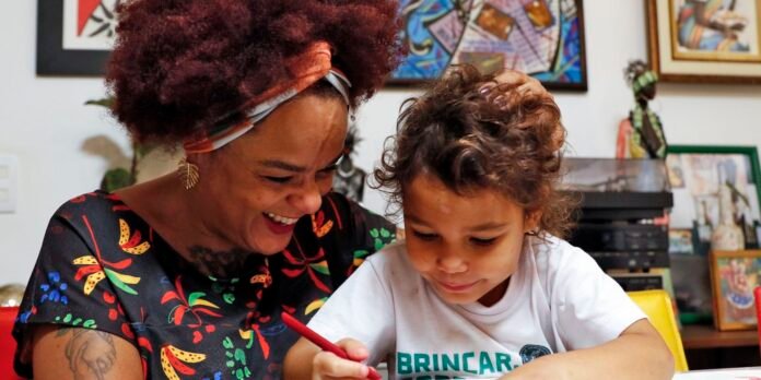 Smiling woman with curly red hair and headband helping a young girl color at a table; colorful artworks appear in the background.