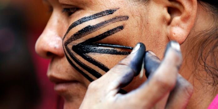 Close-up of a person having bold black geometric face paint applied to the cheek by a makeup artist holding a brush.