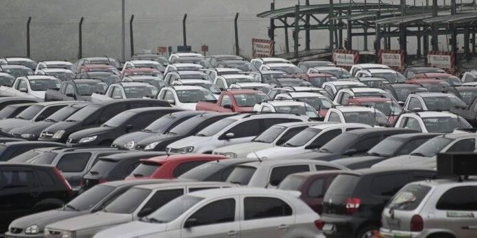 Dense parking lot with hundreds of cars parked closely together under a cloudy sky, behind a chain-link fence and structure in the background.