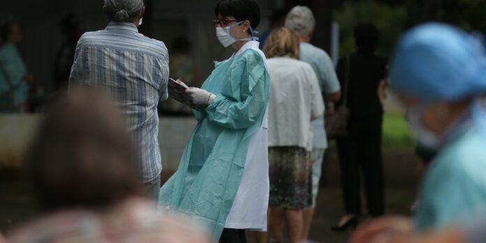 Healthcare worker in teal protective gown and mask checks a clipboard while people stand in line outdoors.