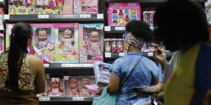 Shoppers browse a toy aisle filled with boxed dolls and pink packaging on store shelves.