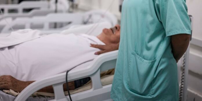 Nurse in teal scrubs stands beside a patient lying in a hospital bed, covered with white sheets.