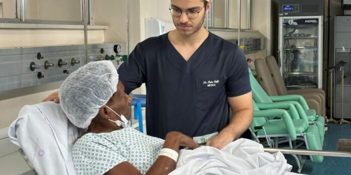 Male healthcare worker in navy scrubs sits beside an elderly patient in a hospital bed, holding hands in a clinical room.