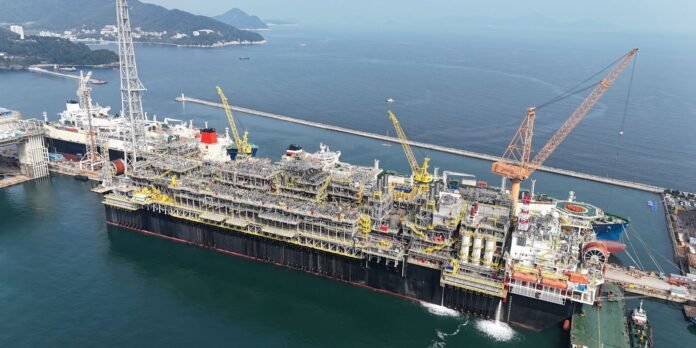 Offshore drilling platform barge docked at a harbor with yellow cranes and complex piping structures nearby.