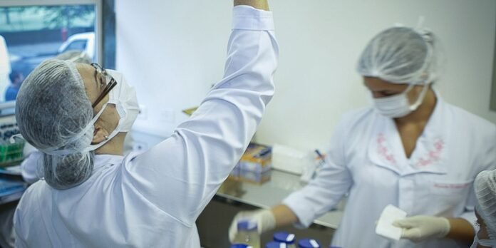 Two scientists in protective gear (white coats, hairnets, masks) work at a lab bench, one raising an arm as they handle supplies.