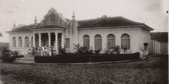 Historic villa with a tiled roof, arched windows, and a columned front porch; a group of people posing on the steps