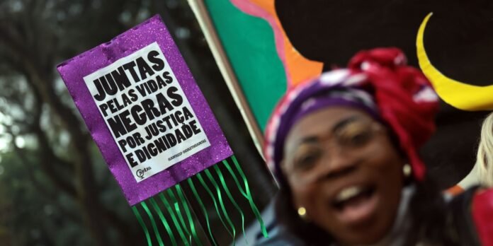 Purple glitter protest sign reading 'Juntas pelas vidas negras por justiça e dignidade' with a smiling person in a colorful headwrap in the background.