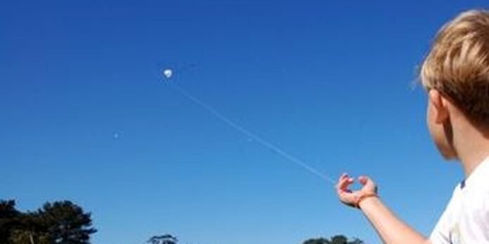 Person with blond hair seen from behind, holding a kite string against a clear blue sky with a small white kite high up.