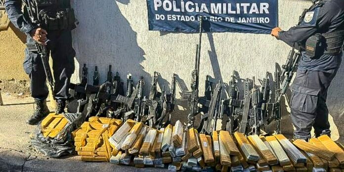 Polícia Militar officers guard a display of seized guns and stacked gold bars laid out on the ground in front of a banner.