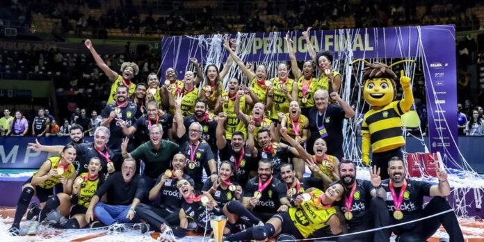 A sports team in yellow and black uniforms poses with medals on a podium, celebrating a victory with confetti and cheers.