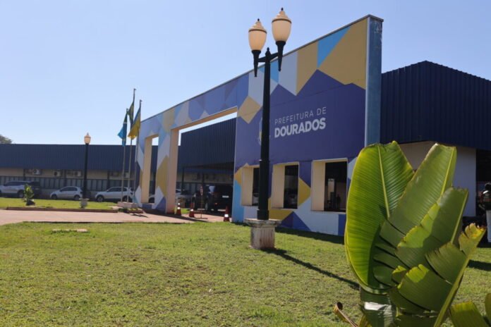 Exterior of Prefeitura de Dourados with a blue-yellow geometric mural, flags, streetlamps, and a large green plant in the foreground.