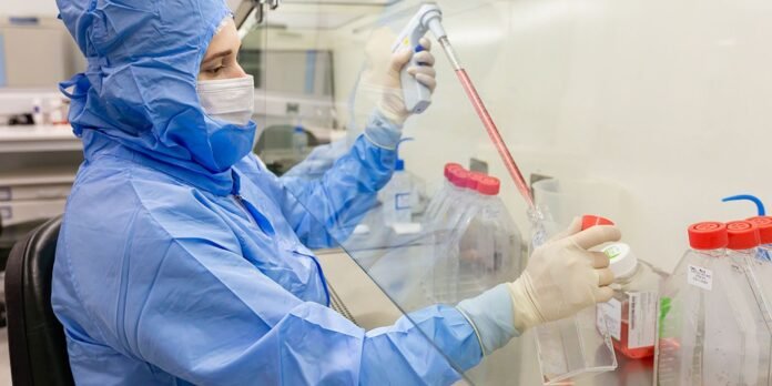 Laboratory technician in blue protective suit and mask uses a pipette inside a biosafety cabinet.