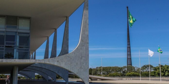Modern concrete building with curved archways and tall flagpoles flying Brazil flags against a clear blue sky.