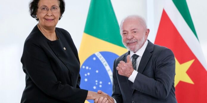 Two officials shake hands and smile, with multiple national flags in the background.