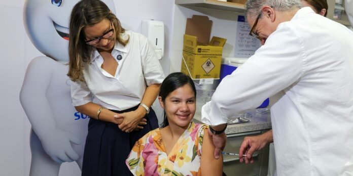 A smiling young woman wearing a floral dress sits as a clinician administers a vaccination in a medical clinic, with staff nearby.