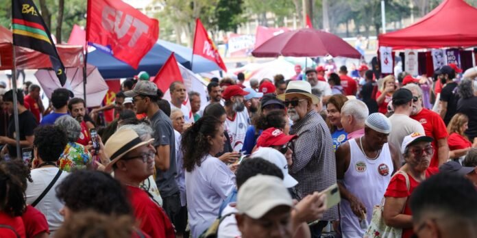 Crowded outdoor market or rally with red flags, tents, and diverse people talking and walking.