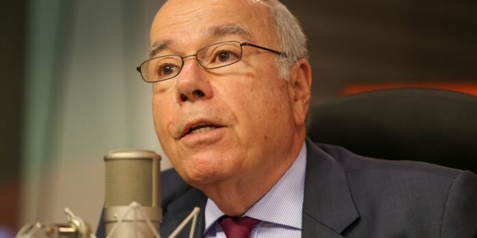 Man in a dark suit and red tie speaking into a microphone in a studio setting, wearing glasses.