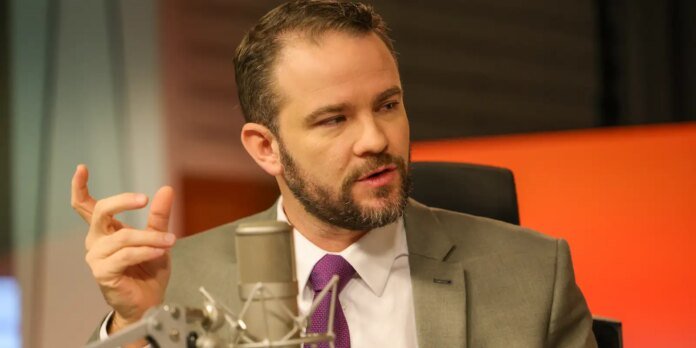 Man in a gray suit and purple tie speaks into a studio microphone during a podcast/recording session, gesturing with his hand.