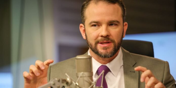 Bearded man in a gray suit and purple tie speaks into a microphone in a recording studio.