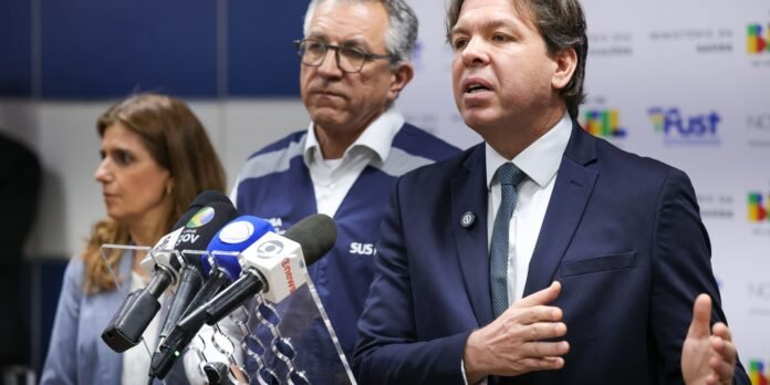 Man in a navy suit speaking at a press conference with multiple microphones in front of a backdrop with logos. Another person stands behind him.