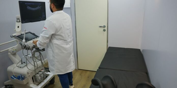 Medical professional in a white coat operating an ultrasound machine in a clinical exam room with a padded bed nearby.