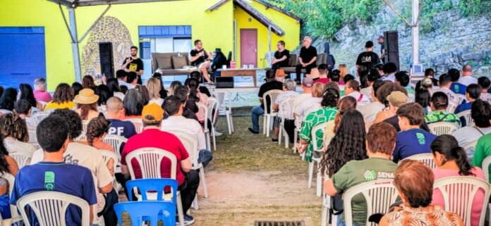 Outdoor panel discussion with five speakers on a stage under a canopy, facing a large seated audience on plastic chairs.