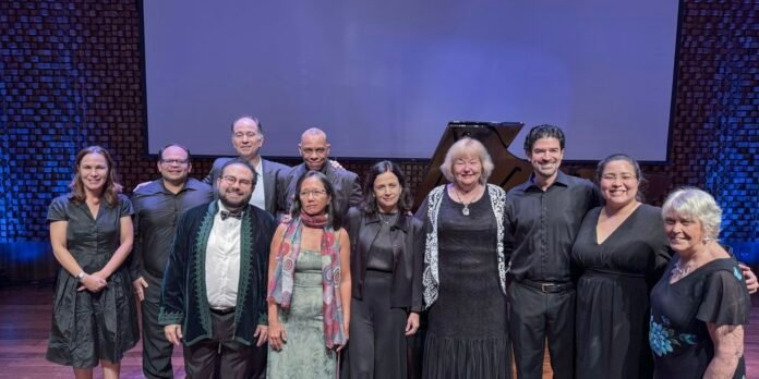 Group of eleven performers posing together on a stage, a grand piano behind them, dressed in formal black attire with one wearing a velvet jacket and scarf edge.