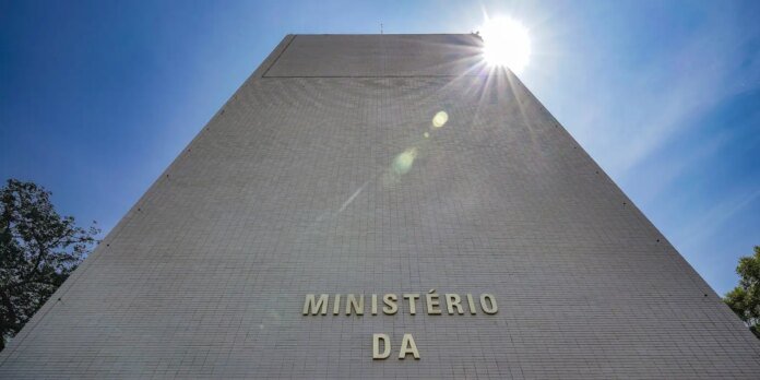 White tiled pyramid-like government building with the inscription 'MINISTÉRIO DA' on the front, bright sun flare at the top.