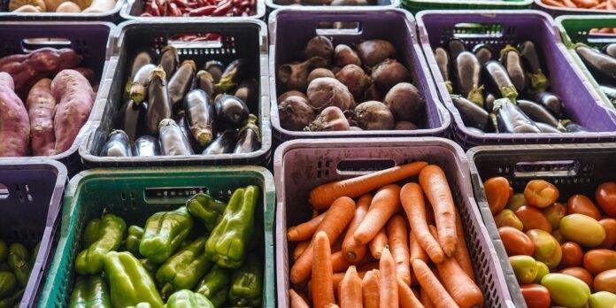 Crates of fresh vegetables at a market: carrots, green peppers, eggplants, potatoes, and tomatoes in colorful boxes.