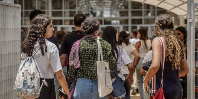 A group of students walking through an outdoor walkway toward a glass building, seen from behind with backpacks and tote bags visible.