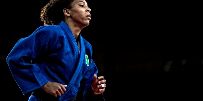 Female judoka in a blue gi running or moving forward, focused profile view against a dark background. Patch on gi visible.