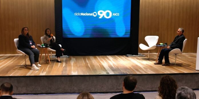Panel discussion on a stage with a large blue screen reading 