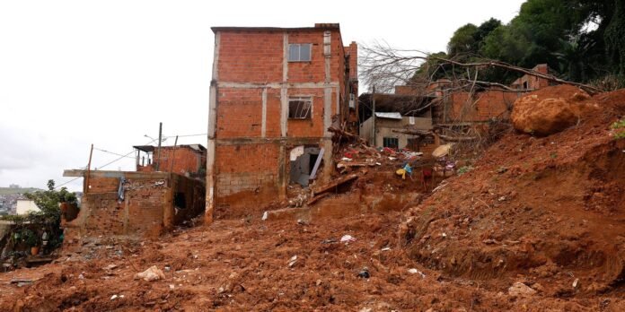 Red brick buildings on a muddy, debris-strewn hillside with collapsed walls and scattered trash; a fallen tree lies across the scene.