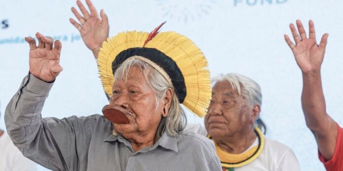 Elderly Indigenous man in a wide yellow feather headdress raises his hand on stage during a cultural performance.