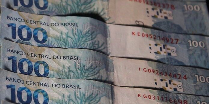 Stacks of Brazilian 100 reais banknotes with 'Banco Central do Brasil' and the number 100 visible, blue-toned design.