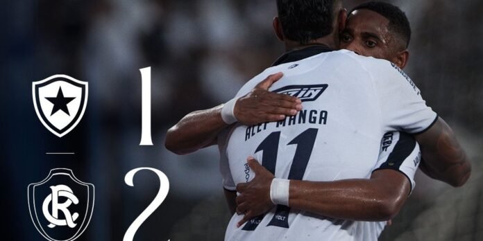 Two soccer players embrace in celebration as the scoreboard shows 1–2, with team crests on the left side.