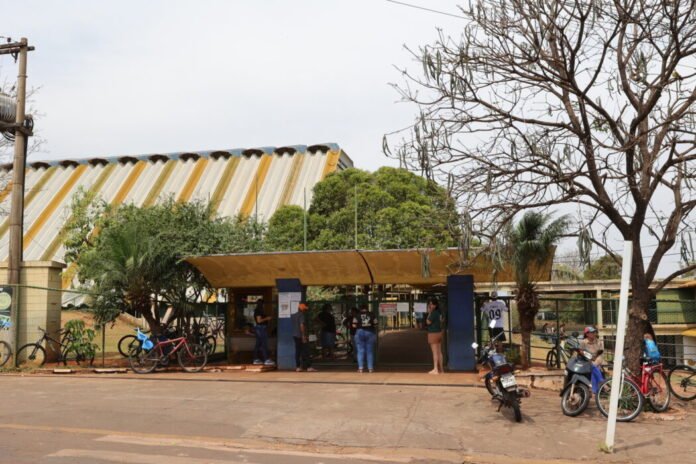 Open-air shelter with a yellow roof where people stand and sit, bikes and motorcycles parked nearby on a sunny street.
