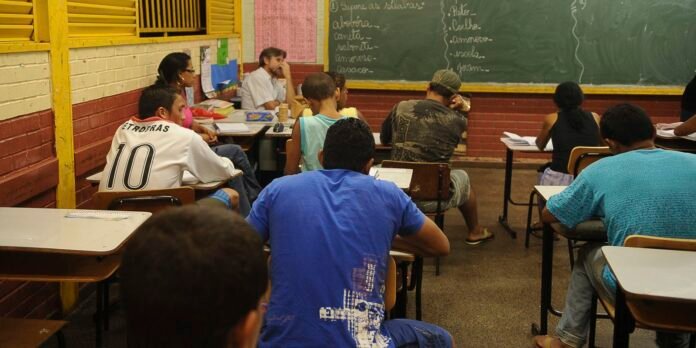 Diverse group of students studying in a classroom; desks in rows, chalkboard with notes at the front, brick walls and yellow window shutters.