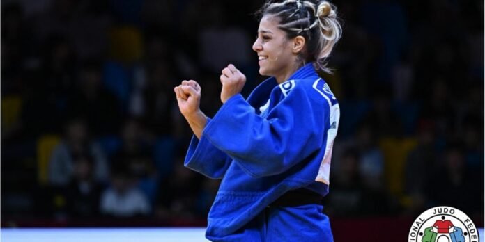 Female judoka in a blue gi raising fists in celebration with a crowd in the background and the IJF logo visible in the corner.