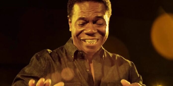 Smiling man in a dark, warm-lit setting with hands raised as if gesturing or performing on stage
