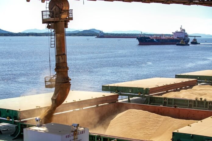 Industrial port scene with a large crane feeding a conveyor that dumps sand onto stacked piles, ships visible in the water.