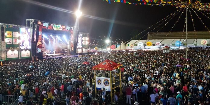 Huge crowd at a nighttime outdoor concert in front of a brightly lit stage with screens and festival banners.