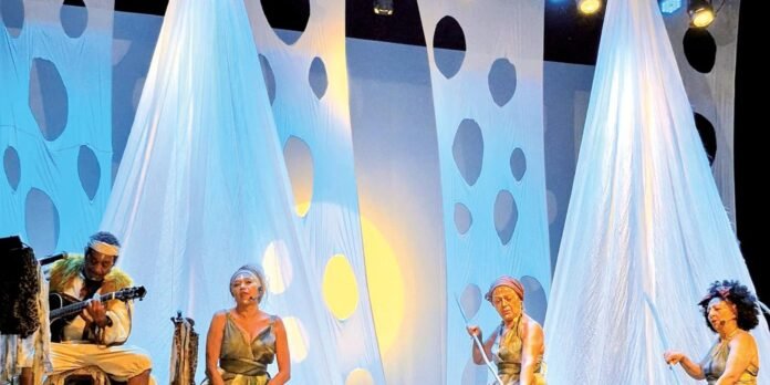 Stage performance with four musicians in gold costumes; a guitarist on the left and three women seated among billowing blue fabric panels with circular cutouts, under warm stage lighting.
